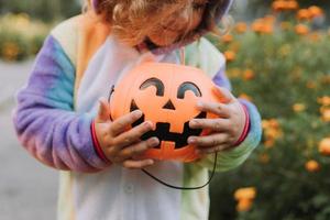 süßes kleines Mädchen in einem Regenbogen-Einhorn-Kostüm für Halloween geht, um Süßigkeiten in einem Kürbiskorb in einem Wohngebiet zu sammeln. Kind geht im Freien spazieren. Süßes oder Saures. Lebensstil. Kigurumi foto