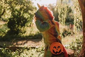 Kleines Mädchen in einem Regenbogen-Einhorn-Halloween-Kostüm und ein Dackel in einem Kleid mit einem Kürbiskorb für Süßigkeiten sitzen bei Waldsonnenuntergang auf Baumstumpf. fabelhafter wunderbarer Zauberwald. Platz für Text foto