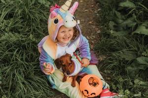 Kleines Mädchen in einem Regenbogen-Einhorn-Halloween-Kostüm und ein Dackel in einem Kleid mit einem Kürbiskorb für Süßigkeiten sitzen bei Waldsonnenuntergang auf Baumstumpf. fabelhafter wunderbarer Zauberwald. Platz für Text foto