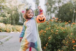 süßes kleines Mädchen in einem Regenbogen-Einhorn-Kostüm für Halloween geht, um Süßigkeiten in einem Kürbiskorb in einem Wohngebiet zu sammeln. Kind geht im Freien spazieren. Süßes oder Saures. Lebensstil. Kigurumi foto