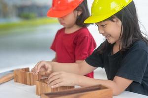 asiatische geschwistermädchen, die technikhüte tragen, bauen häuser aus dem holzspielzeug. um zu lernen und die Entwicklung zu verbessern, kleiner Architekt. foto