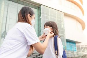 asiatische mutter hilft ihrer tochter, die eine medizinische maske zum schutz von covid-19 oder coronavirus-ausbruch trägt, sich darauf vorzubereiten, zur schule zu gehen, wenn sie wieder in der schule sind. foto