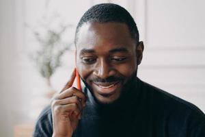 fröhlicher junger afroamerikanischer geschäftsmann, der auf dem smartphone spricht, während er fern zu hause arbeitet foto