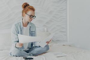 Junge konzentrierte Hausfrau mit Brille, die das Familienbudget verwaltet, während sie zu Hause auf dem Bett sitzt foto