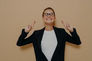 junge hoffnungsvolle frau mit brille in schwarzem blazer über weißem t-shirt, die hoffnungsvoll die daumen drückt foto