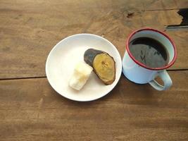 eine Tasse schwarzer Kaffee, gekochter Maniok und gekochte Banane auf einem Tisch aus Holzbrettern foto