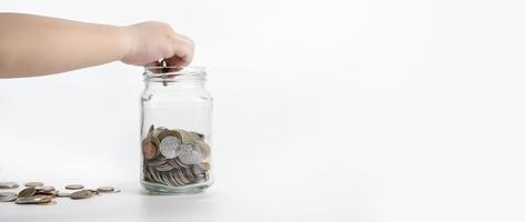 Die Hand des Kindes steckt eine Münze in ein Glasgefäß, das auf weißem Hintergrund isoliert ist. geldsparkonzept, finanzielle geschäftsinvestitionen, münze in glasflasche foto