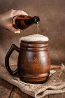 Bierlehm brauner Krug mit Bier auf einem Holztisch auf dunklem Hintergrund. ein strahl helles bier ergießt sich in ein glas foto