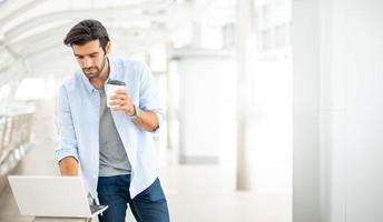 junger kaukasischer mann, der eine tasse kaffee hält und an einem freiberuflichen projekt mit einem tragbaren computer im äußeren büro arbeitet. foto