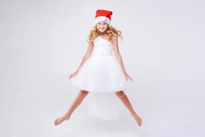 kleines fröhliches mädchen in roter weihnachtsmütze und weißem schönem kleid springt ab foto