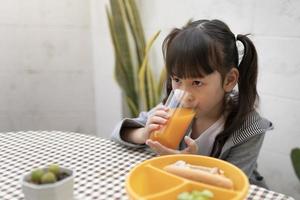 Schönes junges Mädchen, das zu Hause Orangensaft und Frühstück auf dem Tisch trinkt. kind glücklich essen, fruchtsaft, gesunde ernährung, essen und snacks foto