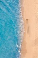 Luftaufnahme eines Paares, das am Strand mit Sonnenlicht in der Nähe von türkisfarbenen Meereswellen spazieren geht. draufsicht auf die sommerstrandlandschaft, romantischer inspirierender paarurlaub, romantikurlaub. Vorlage für Freiheitsreisen foto