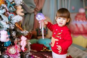 Porträt eines kleinen Mädchens im roten Weihnachtspullover steht am Weihnachtsbaum foto