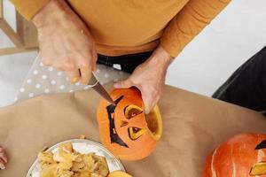 Nahaufnahme von Mannhänden, die Jack o Laternen aus reifem orangefarbenem Kürbis mit Messer auf ihrem hölzernen Küchentisch schnitzen. mann, der all hallows eve halloween partydekorationen vorbereitet. Hintergrund, Kopierbereich. foto