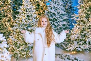 Süßes kaukasisches blondes Mädchen in weißem Öko-Pelzmantel, das im Winterweihnachtswald mit Lichtern spaziert. neues Jahr, Märchenkonzept foto