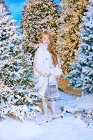 Süßes kaukasisches blondes Mädchen in weißem Öko-Pelzmantel, das im Winterweihnachtswald mit Lichtern spaziert. neues Jahr, Märchenkonzept foto