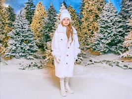 Süßes kaukasisches blondes Mädchen in weißem Öko-Pelzmantel, Hut und Handschuhen, das im Winterweihnachtswald spazieren geht. neues jahr, märchen, modekonzept foto