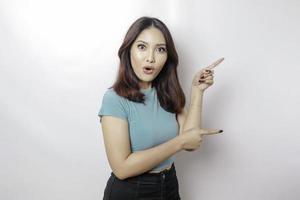 schockierte asiatische Frau mit blauem T-Shirt, die auf den Kopierbereich neben ihr zeigt, isoliert durch weißen Hintergrund foto