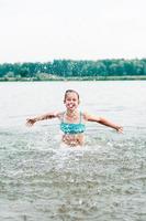 Ein fröhliches Mädchen steht mit ausgestreckten Armen hinter den Spritzern im Fluss. lokaler Tourismus foto