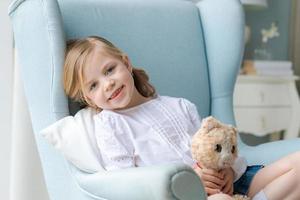 süßes kaukasisches mädchen sitzt zu hause im schlafzimmer in einem blauen sessel am fenster foto