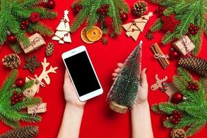 Blick von oben auf die weiblichen Hände, die das Telefon in der einen Hand und den Weihnachtsbaum in der anderen Hand auf rotem Hintergrund halten. neujahrsferienkonzept. Attrappe, Lehrmodell, Simulation foto