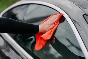 bemannt die hand mit lappen, der seitenfenster des schwarzen autos reinigt. professionelles autowaschkonzept. regelmäßiges Abwaschen. Abwischen von Wassertropfen am Fahrzeug foto