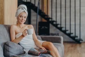 Das Foto einer entzückenden jungen Frau hat Make-up und gesunde Haut, hält eine Tasse Kaffee in der Hand, ist in ein Badetuch gewickelt, liest eine Zeitschrift und fühlt sich entspannt auf dem Sofa, verbringt Freizeit zu Hause. Trost empfinden