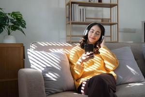 junge asiatin, die musik über kopfhörer hört und eine notiz für ihre arbeitsidee schreibt. sie sitzt auf einem grauen sofa im wohnzimmer. foto