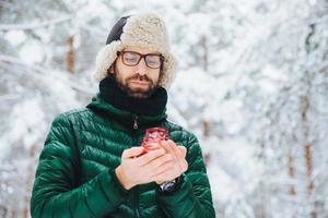 attraktiver ernster mann trägt warme winterkleidung, hält kerze, steht vor winterwaldhintergrund, genießt frische luft, ist sehr selbstbewusst. Modischer unrasierter Mann im Freien. foto