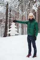 vertikales porträt eines positiv lächelnden mannes trägt modische warme kleidung, hält weiße tanne in den händen, posiert im weißen winterwald. glücklicher unrasierter attraktiver mann verbringt freizeit oder winterferien foto