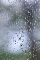 ein Foto von Regentropfen auf dem Fensterglas mit verschwommenem Blick auf die blühenden grünen Bäume. abstraktes Bild, das bewölkte und regnerische Wetterbedingungen zeigt