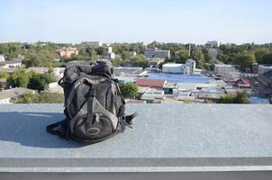 Der schwarze Rucksack liegt bei sonnigem Wetter auf dem Metallrand des Daches eines mehrstöckigen Wohngebäudes foto