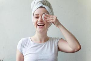 Lächelnde Frau im Handtuch auf dem Kopf mit weicher, gesunder Haut, die das Make-up mit einem Wattepad entfernt, das auf weißem Hintergrund isoliert ist foto