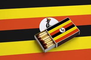 Die Uganda-Flagge wird in einer offenen Streichholzschachtel gezeigt, die mit Streichhölzern gefüllt ist und auf einer großen Flagge liegt foto