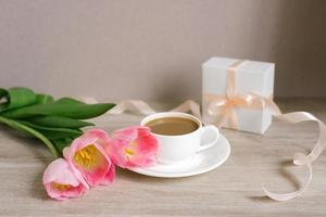 Kaffee mit Milch in einer weißen Porzellantasse und Untertasse, ein Strauß frühlingsrosa Tulpen und ein Geschenk in einer weißen Schachtel mit Satinband. Muttertag, Valentinstag, Ostern. das Konzept des Frühlings foto