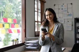 lächelnde schöne asiatische geschäftsfrau, die diagramm und diagramm analysiert, die änderungen auf dem markt zeigen und smartphone im büro halten. foto