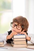 müder Schuljunge. Süßer kleiner Junge, der schläft, während er am Tisch sitzt und sein Gesicht am Bücherstapel lehnt foto