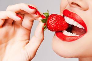 Frische kommt in Rot. Nahaufnahme einer schönen Frau, die Erdbeere in der Hand hält und sie schmeckt, während sie vor grauem Hintergrund steht foto