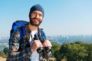 gute Kulisse. hübscher junger mann, der rucksack trägt und mit einem lächeln in die kamera schaut, während er in der natur steht und ein fernglas hält foto