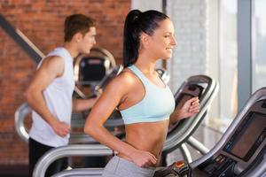 Cardio Training. Seitenansicht einer schönen jungen Frau, die auf einem Laufband läuft und mit einem Mann im Hintergrund lächelt foto