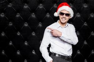 Schauen Sie sich diesen hübschen jungen Mann mit Sonnenbrille und Weihnachtsmütze an, der wegzeigt und lächelt, während er vor schwarzem Hintergrund steht foto