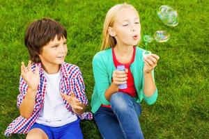 Blasenspaß. Zwei süße kleine Kinder, die Seifenblasen blasen, während sie zusammen auf dem grünen Gras sitzen foto