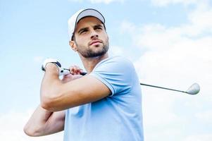 perfekter Schlag. Tiefwinkelansicht des jungen Golfers, der seinen Fahrer schwingt und mit blauem Himmel als Hintergrund wegschaut foto