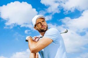 professioneller Schwung. Tiefwinkelansicht des jungen und selbstbewussten Golfers, der seinen Fahrer schwingt und mit blauem Himmel als Hintergrund wegschaut foto