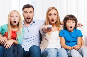 das ist einfach erstaunlich überraschte vierköpfige Familie, die sich aneinander bindet und den Mund offen hält, während sie zu Hause zusammen fernsieht foto