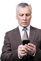 Geschäftstechnologien. Selbstbewusster, reifer Mann in Abendkleidung, der ein Handy hält und es betrachtet, während er vor weißem Hintergrund steht foto