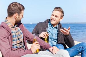 sich Zeit nehmen, um mit einem Freund zu sprechen. Zwei hübsche junge Männer trinken Bier und reden miteinander, während sie zusammen am Strand sitzen foto