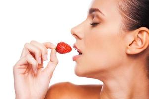Essen mit Leidenschaft. Seitenansicht einer schönen jungen Frau mit nacktem Oberkörper, die eine Erdbeere in der Hand hält und die Augen geschlossen hält, während sie vor weißem Hintergrund steht foto