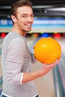 auf einen Streik hoffen. Fröhlicher junger Mann, der über die Schultern schaut und eine Bowlingkugel hält, während er gegen Bowlingbahnen steht foto