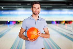 Bowlingmeister. Fröhlicher junger Mann, der eine Bowlingkugel hält und in die Kamera lächelt, während er gegen Bowlingbahnen steht foto